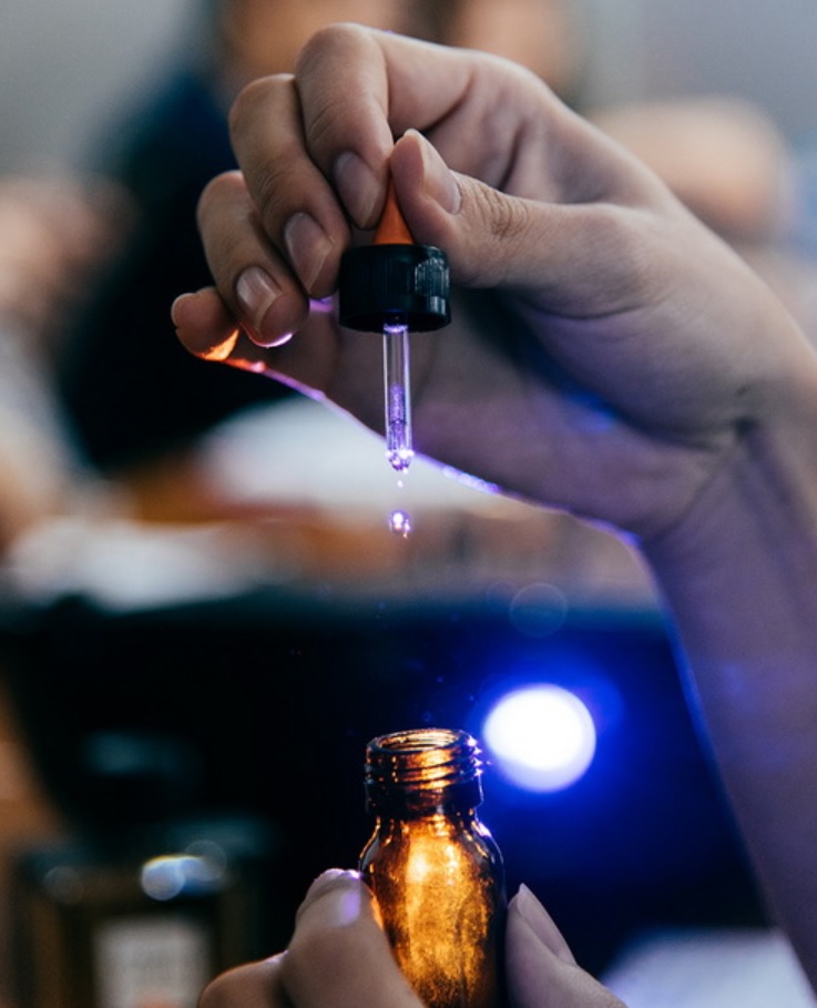Hands holding a glass dropper above an amber material bottle as a drop falls.